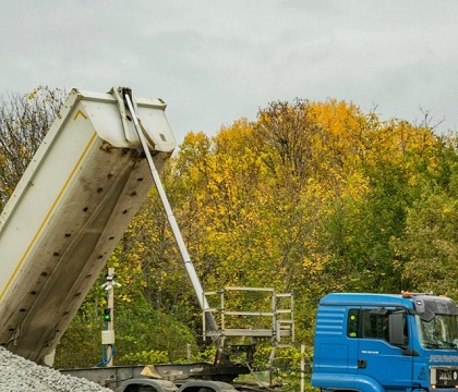 A blue Khon Structure tipper truck with its dump body raised, parked on gravel beside autumn trees