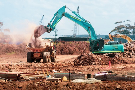 A teal tracked excavator loading red earth into an articulated dump truck at a construction earthworks site.